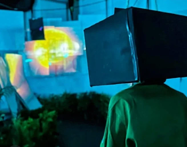 A visitor with a box over their head inside the Under25 Summit installation, lit in blue and green